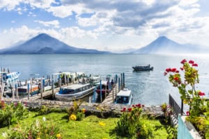 Boats docked at a lake with lush greenery, vibrant flowers, and a scenic volcanic backdrop, showcasing a picturesque lakeside view in Guatemala.