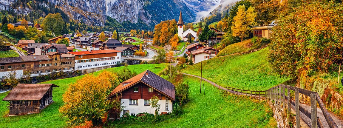 Vibrant autumn scene in a Swiss alpine village nestled in a lush valley surrounded by tall mountains and waterfalls, showcasing scenic landscape, traditional architecture, and colorful fall foliage.