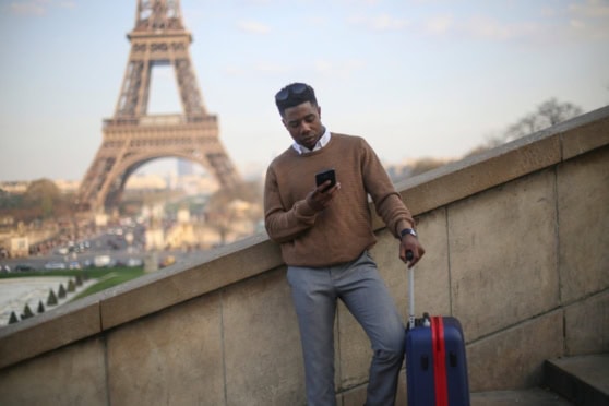 A young man with suitcase using smartphone in Paris with Eiffel Tower in background.