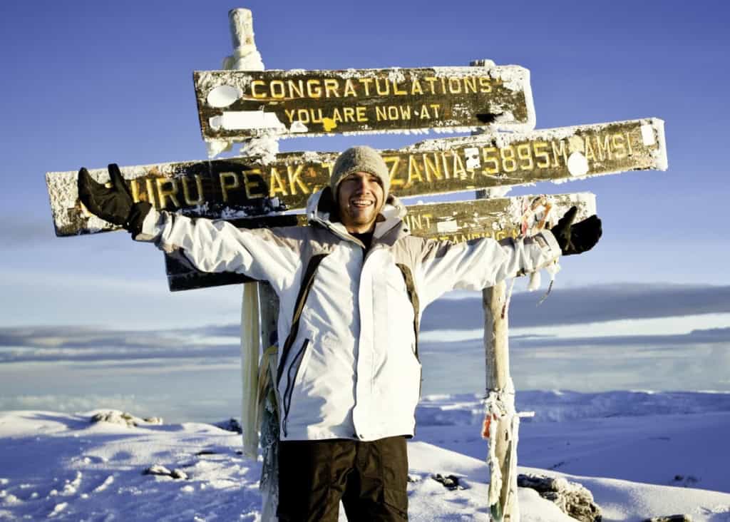 2. Smiling man at Mount Kilimanjaro summit, snowy scenery, clear blue sky, celebrating achievement, outdoor adventure, travel, high-altitude expedition, mountainous terrain, success, outdoor activity.