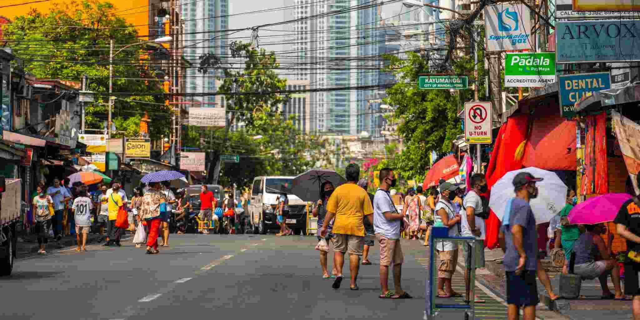 A busy city street scene with diverse pedestrians wearing masks, using umbrellas, and shopping, surrounded by shops and high-rise buildings.