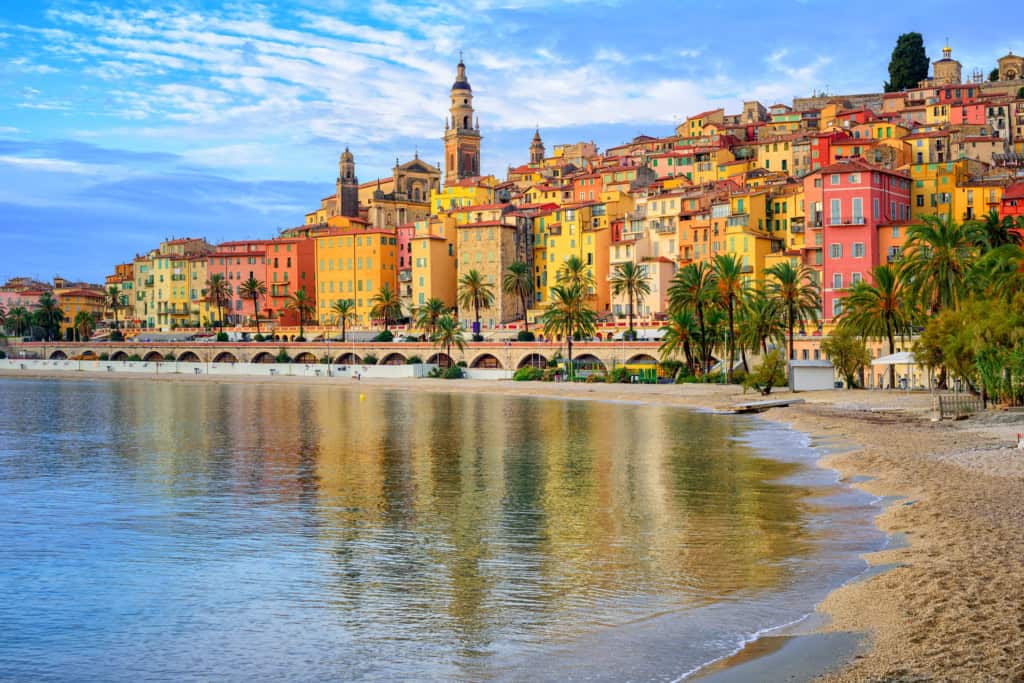 Colorful Mediterranean seaside town with vibrant buildings, palm trees, and historic churches reflected in calm water, showcasing coastal charm and architecture.