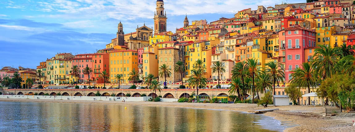 Colorful Mediterranean seaside town with vibrant buildings, palm trees, and historic churches reflected in calm water, showcasing coastal charm and architecture.