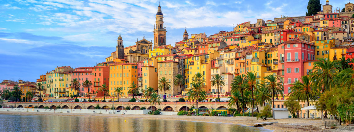 Colorful Mediterranean seaside town with vibrant buildings, palm trees, and historic churches reflected in calm water, showcasing coastal charm and architecture.