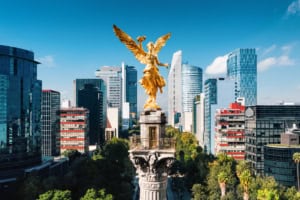 Golden Angel of Independence monument in Mexico City skyline, surrounded by modern glass skyscrapers and lush green trees, representing Mexico's freedom and heritage.