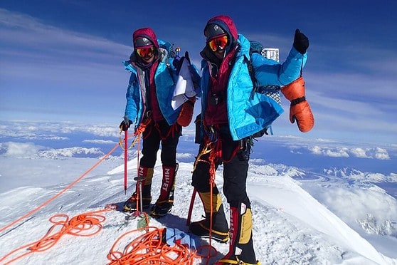 An image of two climbers triumphantly reaching the mountain peak, equipped with safety gear, against a stunning backdrop of snow and sky.