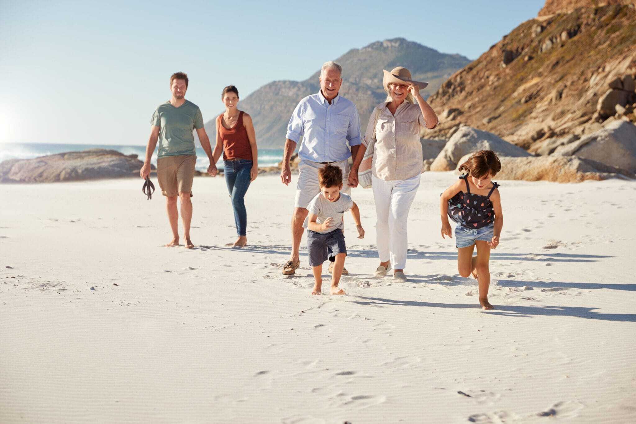 Family enjoying sunny beach day, walking barefoot on sand with mountains nearby, demonstrating the importance of international health cover for travelers in scenic locations.