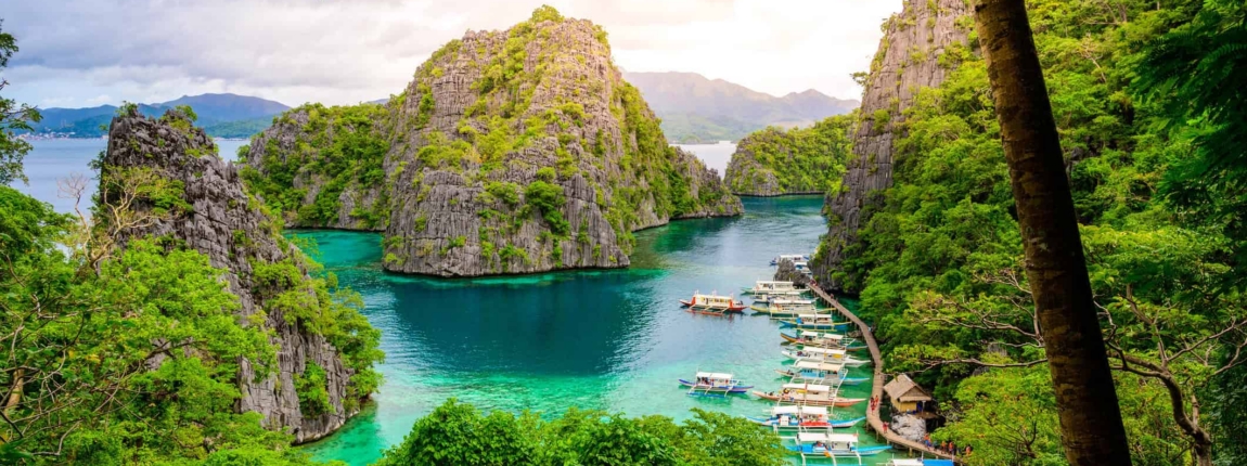Lush tropical island with towering limestone cliffs surrounding a vibrant turquoise lagoon, featuring anchored boats and dense green foliage, representing scenic travel destinations for ICI 2025.
