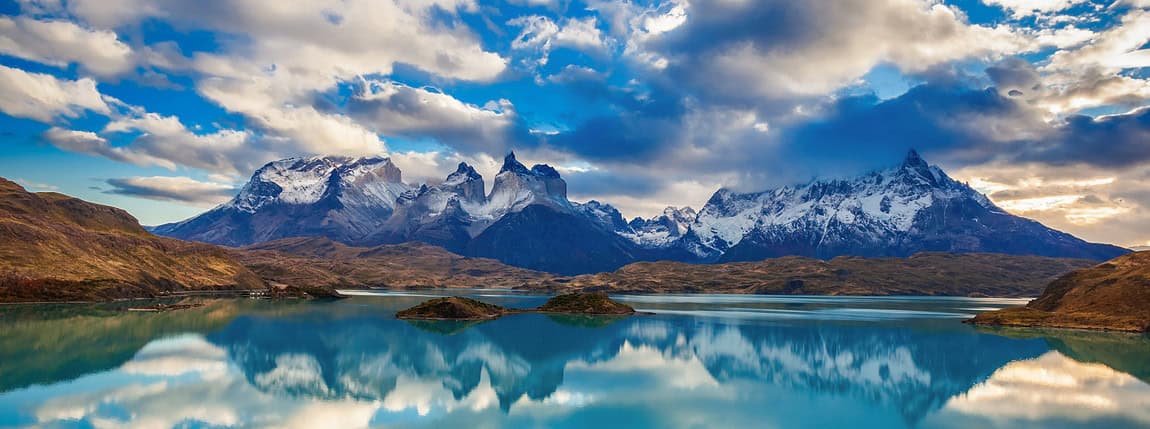 Lush mountain landscape with snow-capped peaks, reflecting on a calm turquoise lake under a dramatic sky in Torres del Paine National Park, Chile. A breathtaking view of Patagonia's natural beauty and wilderness.