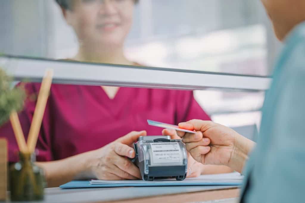 A customer pays at a reception desk with a credit card, with the background showing an insurance agent processing the payment for international health coverage.
