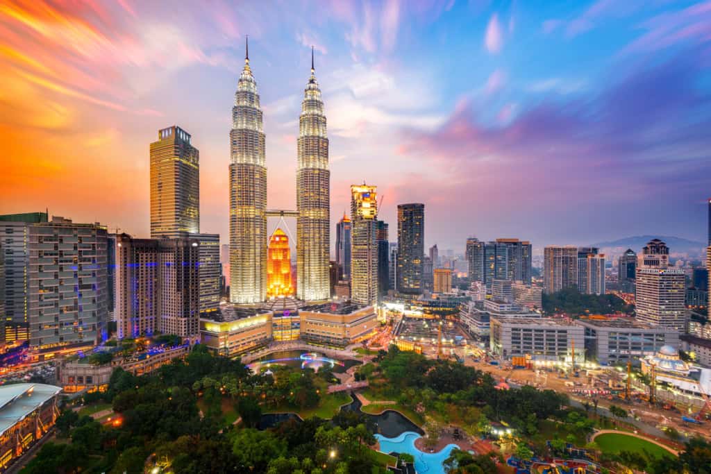 Aerial view of Kuala Lumpur featuring the Petronas Towers illuminated at sunset, with surrounding skyscrapers, lush parks, and busy streets, emphasizing global connectivity and city infrastructure.