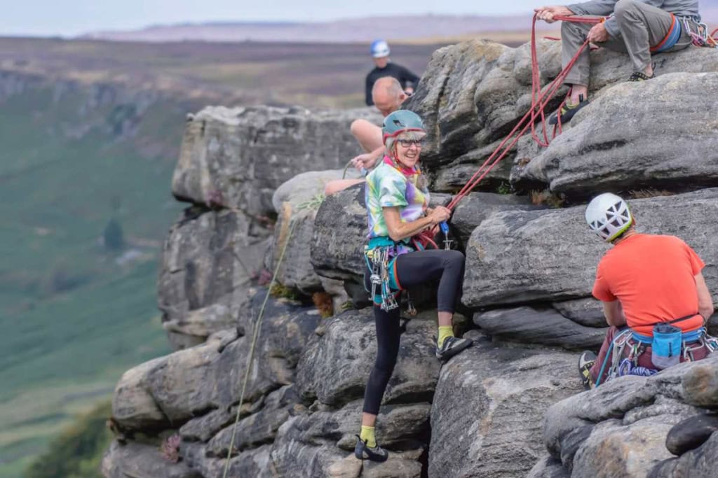 Happy woman climbing rocks with safety gear and a smile, surrounded by friends on a scenic outdoor adventure, showcasing outdoor activity insurance coverage.