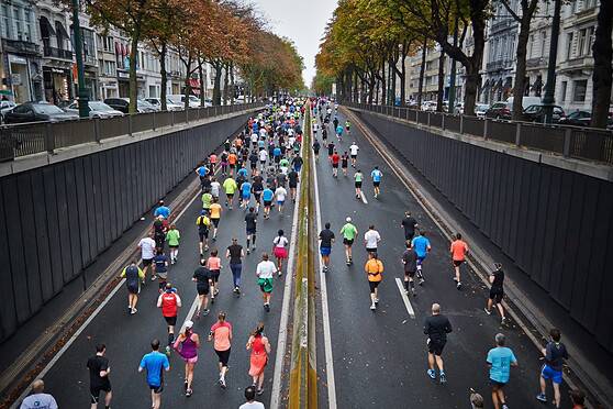 Crowd of runners participating in an urban marathon on a city street lined with trees and historic buildings. The race features diverse participants enjoying outdoor fitness activity.