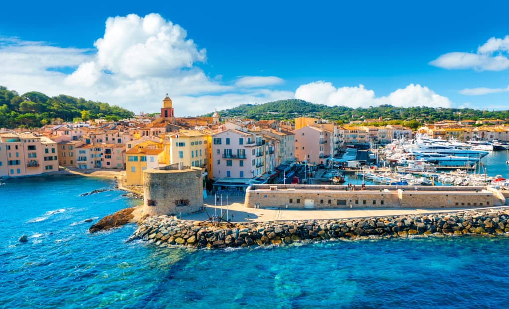 Colorful Mediterranean coastal town with pastel buildings, a marina filled with yachts, historic fortress wall, and lush hills in the background under a bright blue sky.