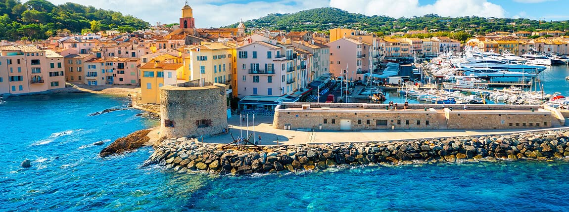 Colorful Mediterranean coastal town with pastel buildings, a marina filled with yachts, historic fortress wall, and lush hills in the background under a bright blue sky.
