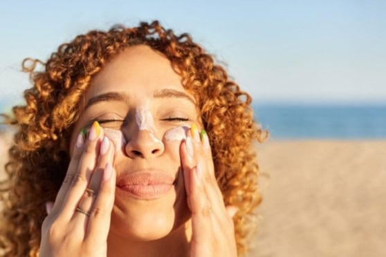 A woman with curly red hair applies sunscreen on her face at the beach, enjoying sunny weather to protect her skin from UV rays.