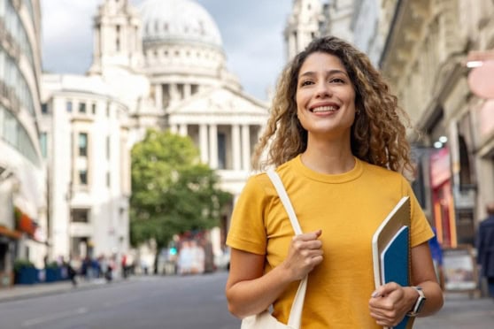 Smiling woman holding notebooks and backpack on city street, representing international travel and insurance needs for global citizens.