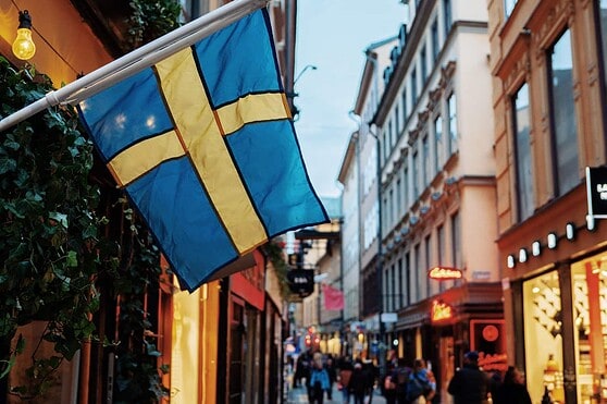A Swedish flag displayed outside a shop on a busy, picturesque city street filled with shops, pedestrians, and charming architecture, illustrating international living and travel opportunities.