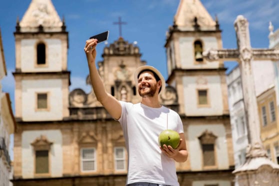 An image of a smiling man taking a selfie in front of a historic European building, holding a green coconut, emphasizing international travel and health insurance for global citizens.