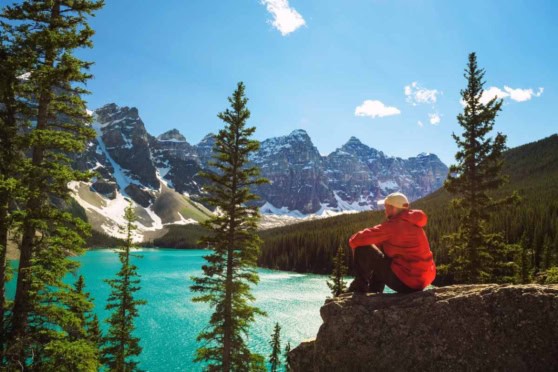 An adventurous hiker enjoying the breathtaking scenery of a mountain lake surrounded by tall pine trees and majestic snow-covered peaks under a bright blue sky.