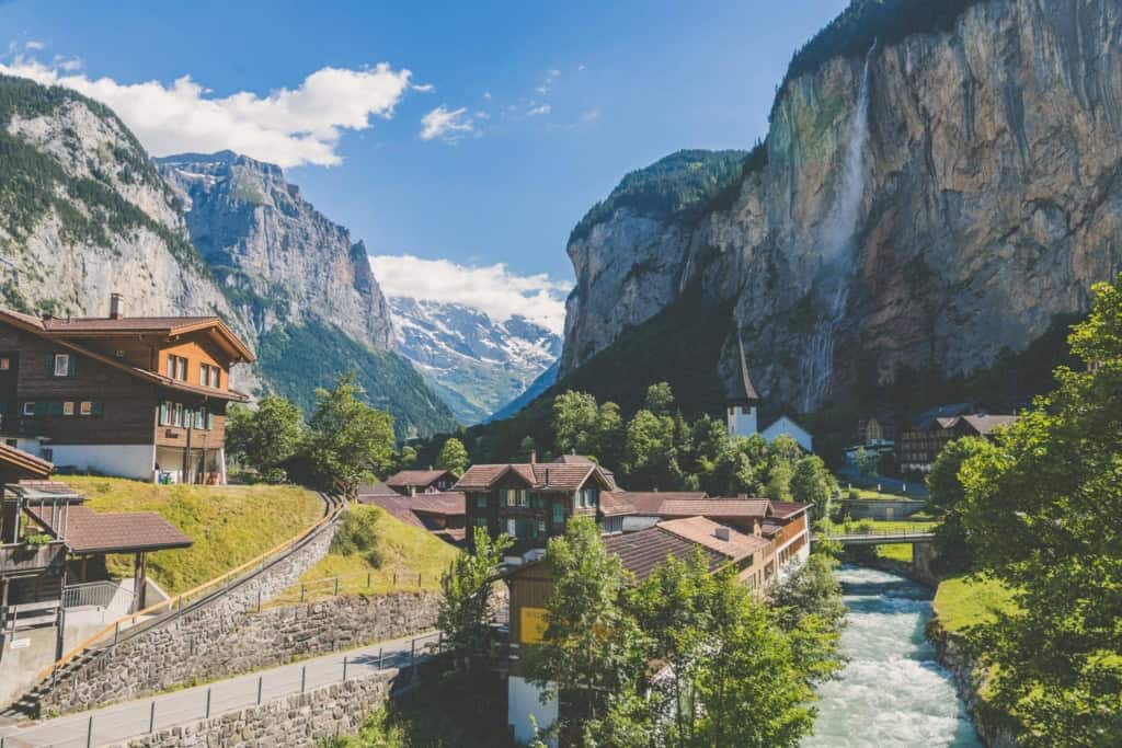 Spectacular Swiss mountain village featuring traditional houses, lush greenery, a flowing river, and majestic waterfalls set against towering Alpine peaks on a sunny day.