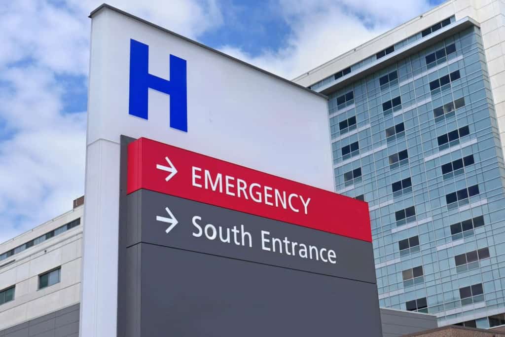 A hospital exterior with a prominent emergency sign indicating the directions to the emergency and south entrance, set against a blue sky with clouds, representing healthcare facilities.