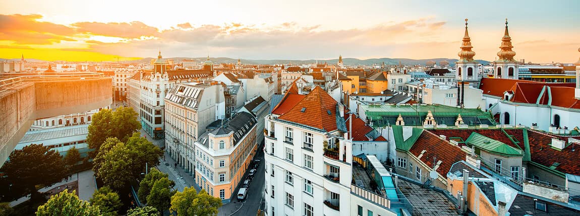Vibrant cityscape of historic European buildings at sunset with orange and pink hues over rooftops and church spires, highlighting urban architecture and scenic views.