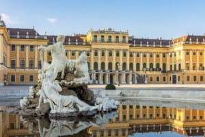 Ornate golden-yellow palace with classical architecture, grand staircase, and lush garden, viewed with a romantic baroque fountain reflection in a serene courtyard, at ICI 2025 event.