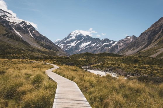 Explore stunning alpine scenery with a winding wooden path leading through lush green fields and snow-topped mountains under a clear blue sky.