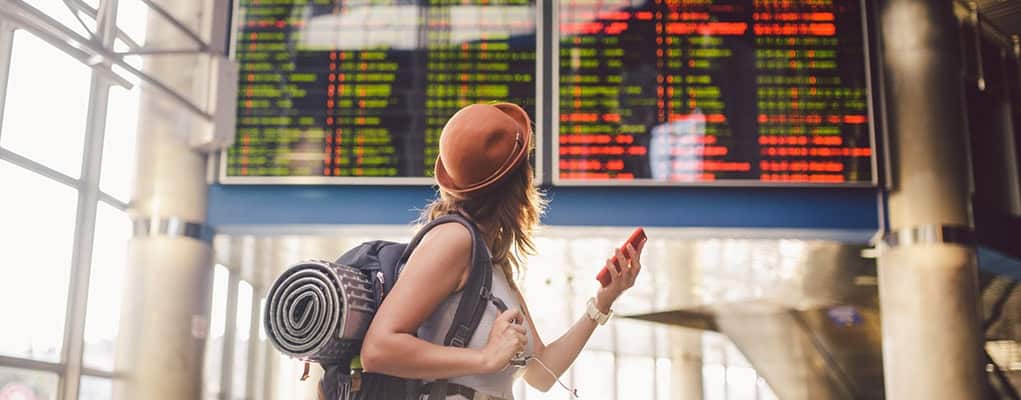 Young woman with backpack checking her phone at airport terminal, standing near flight info display board showing departure and arrival times, biochemistry, and gate details.
