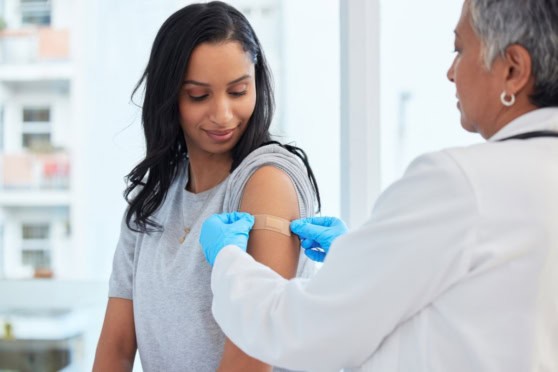 A healthcare professional administering a vaccine shot to a young woman with a bandage on her arm in a clinical setting.