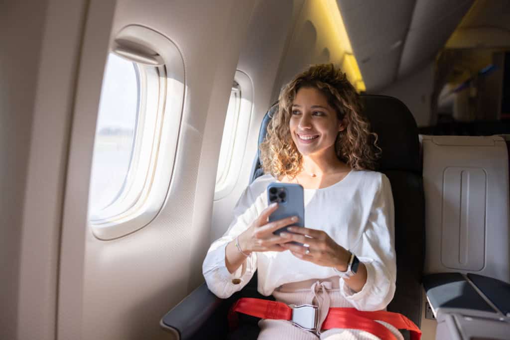 Young woman smiling on airplane, using mobile phone during flight, representing health or travel insurance coverage for international travelers and expats, which is an importance step on a moving abroad checklist