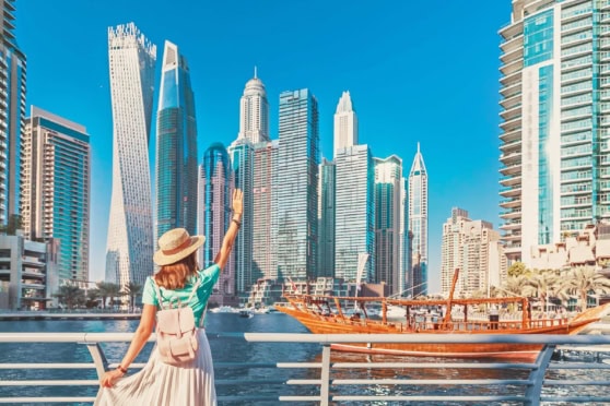 Bright and vibrant Dubai cityscape with high-rise buildings, a woman in summer attire waving, and traditional wooden boat on the water. Suited for topics on international health and travel insurance.