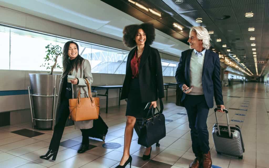 Friendly diverse professionals traveling through airport, smiling with luggage and travel documents, showcasing confidence in international insurance coverage.