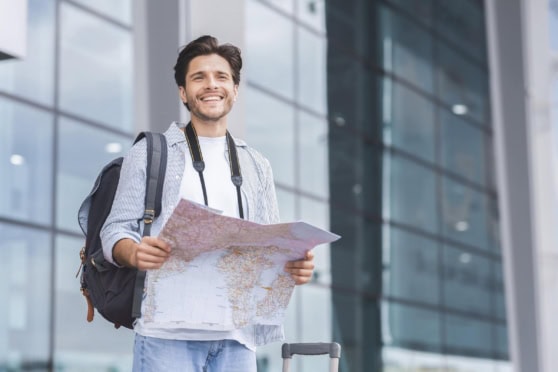 Young man with luggage and map outside modern building, ready for international travel, representing global travel insurance options.