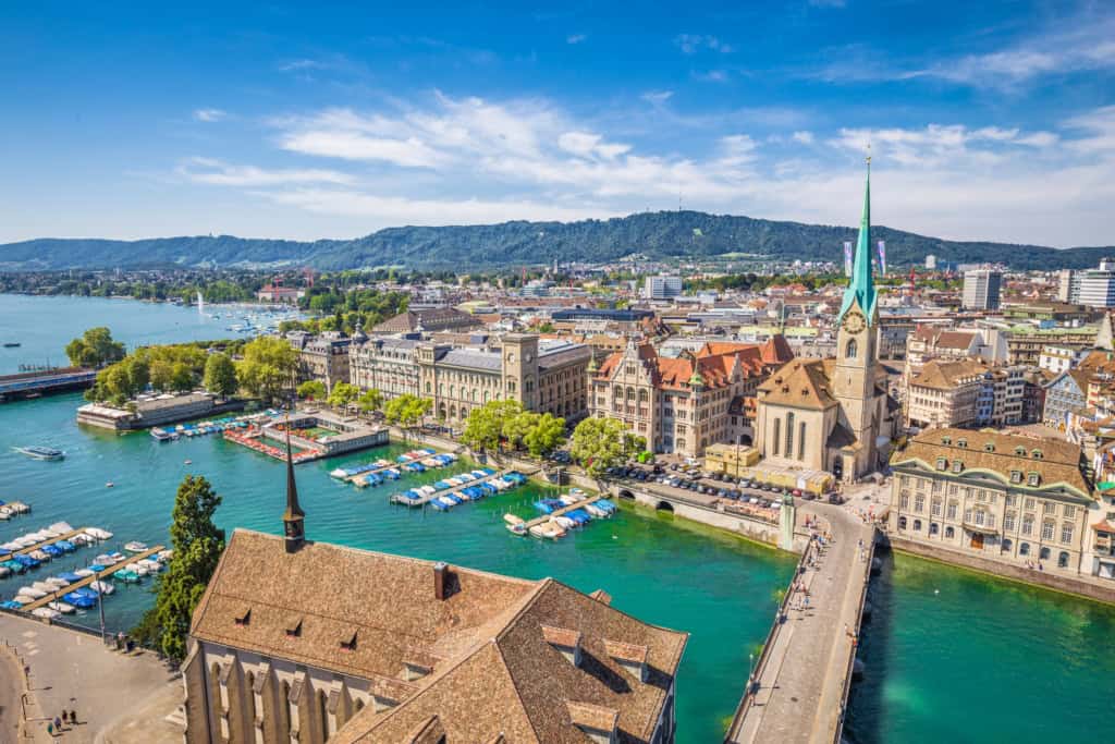 Aerial view of Geneva, Switzerland, showcasing the city skyline, historic churches, and Lake Geneva with boat docks, set against lush green mountains under a bright blue sky.