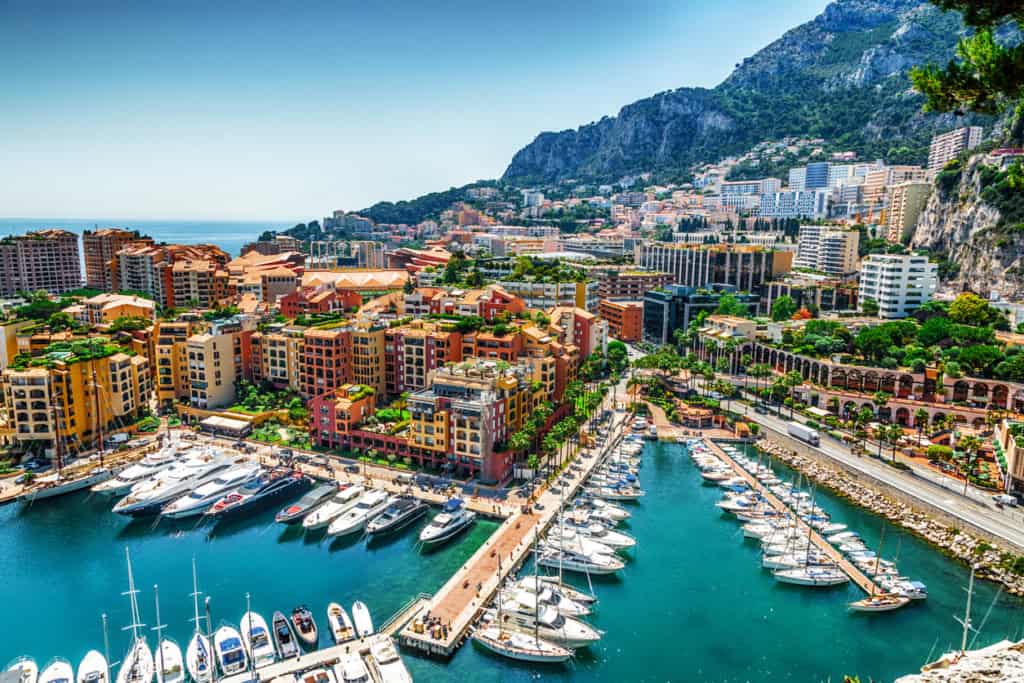 Aerial view of a luxurious marina in a picturesque Mediterranean city, featuring yachts docked along a vibrant waterfront, surrounded by colorful buildings, lush greenery, and stunning mountainous background. Perfect for global citizens in need of reliable international insurance solutions.