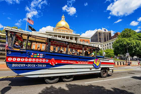 A sightseeing boat on Boston’s Charles River featuring tourists, with historic and modern buildings including the Massachusetts State House and a bright blue sky.
