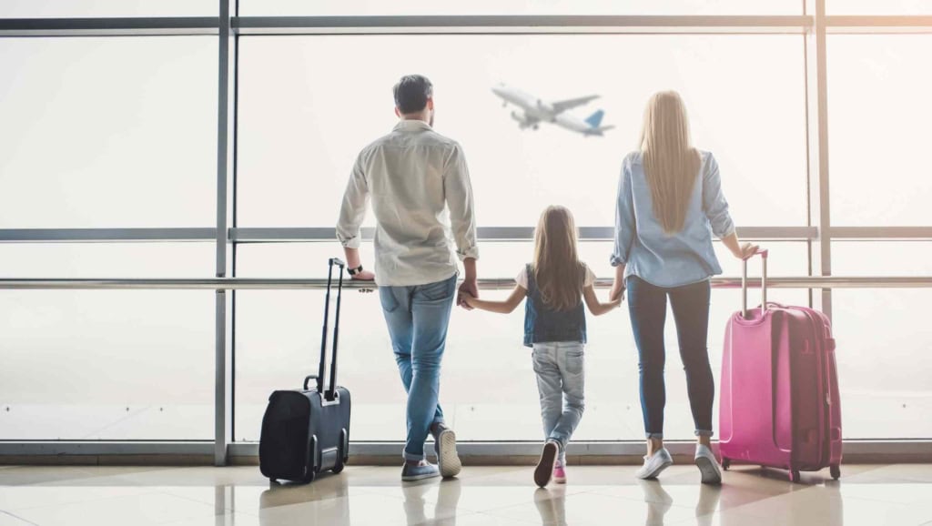 A family with two children at an airport, standing with luggage, watching a plane take off outside the terminal window, representing international travel and insurance coverage.