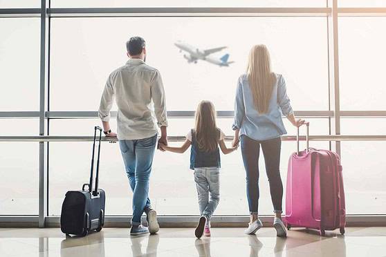 A family with two children at an airport, standing with luggage, watching a plane take off outside the terminal window, representing international travel and insurance coverage.
