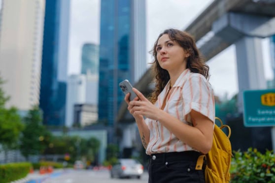 Auto-generated alt text: Young woman with a backpack holding a smartphone, standing in an urban cityscape with high-rise buildings and a metro overhead.