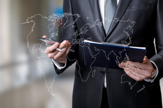 Alt text: Businessman in suit holding a smartphone and notebook with a digital overlay of a world map, representing international insurance services for global citizens.