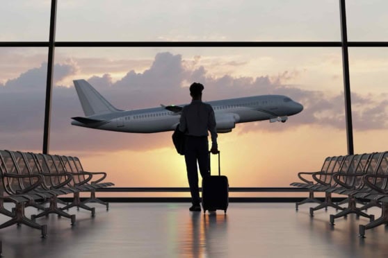 Person waiting at airport terminal with luggage, observing airplane outside window during sunset.