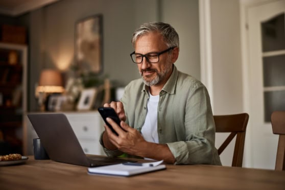 An older man using a mobile phone with a laptop at home, emphasizing international health and insurance solutions for expatriates and travelers worldwide.