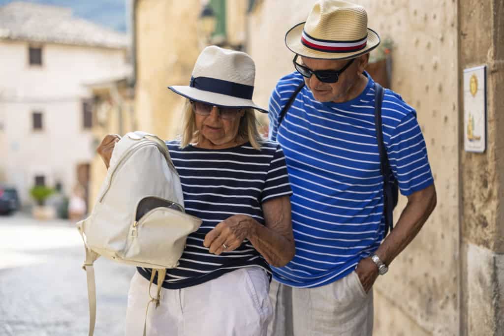 Image of an elderly couple in matching striped shirts and sun hats sightseeing together in a quaint Italian village, with historic architecture in the background.