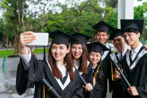 Students in caps and gowns smiling while taking a group selfie at graduation ceremony outdoors.