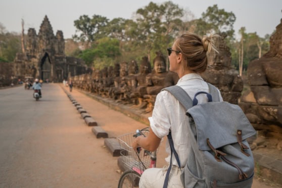 A woman with a backpack and sunglasses exploring an ancient temple site while riding a bicycle, surrounded by lush trees and historical ruins, ideal for travelers seeking adventure and cultural experiences.
