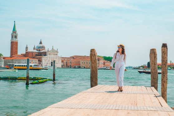 A woman in stylish white attire and sunglasses walks along a Venice dock, showcasing international travel and insurance options.