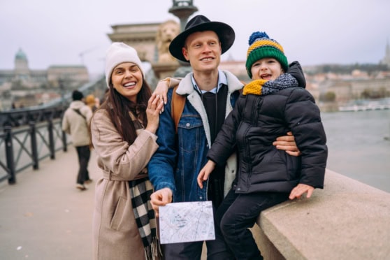 A cheerful family enjoying their trip near a river, dressed warmly in winter clothing, with a scenic cityscape in the background, illustrating protection and peace of mind for international travel.