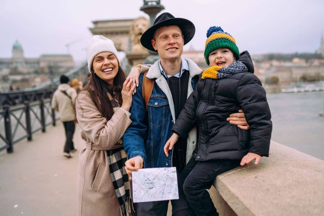 A cheerful family enjoying their trip near a river, dressed warmly in winter clothing, with a scenic cityscape in the background, illustrating protection and peace of mind for international travel.